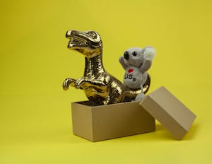 A golden dinosaur and koala toy in an open box with a vibrant yellow backdrop.