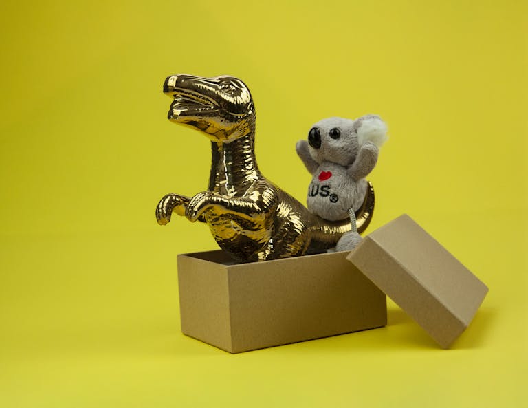 A golden dinosaur and koala toy in an open box with a vibrant yellow backdrop.