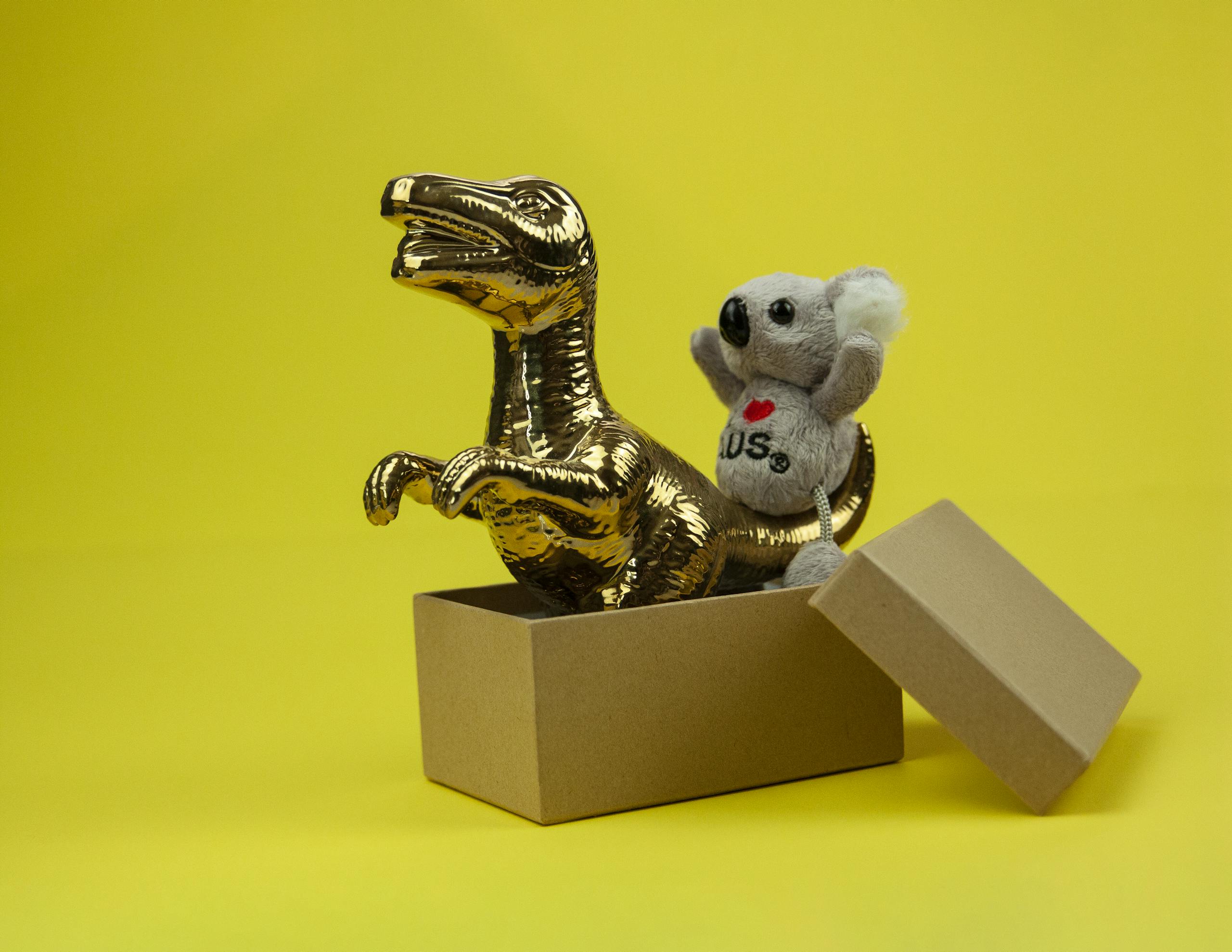 A golden dinosaur and koala toy in an open box with a vibrant yellow backdrop.